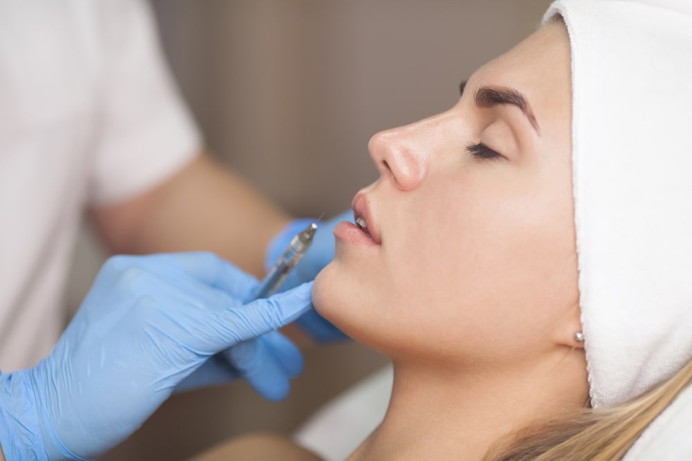 Woman receiving filler injection in lip area by a aesthetician.